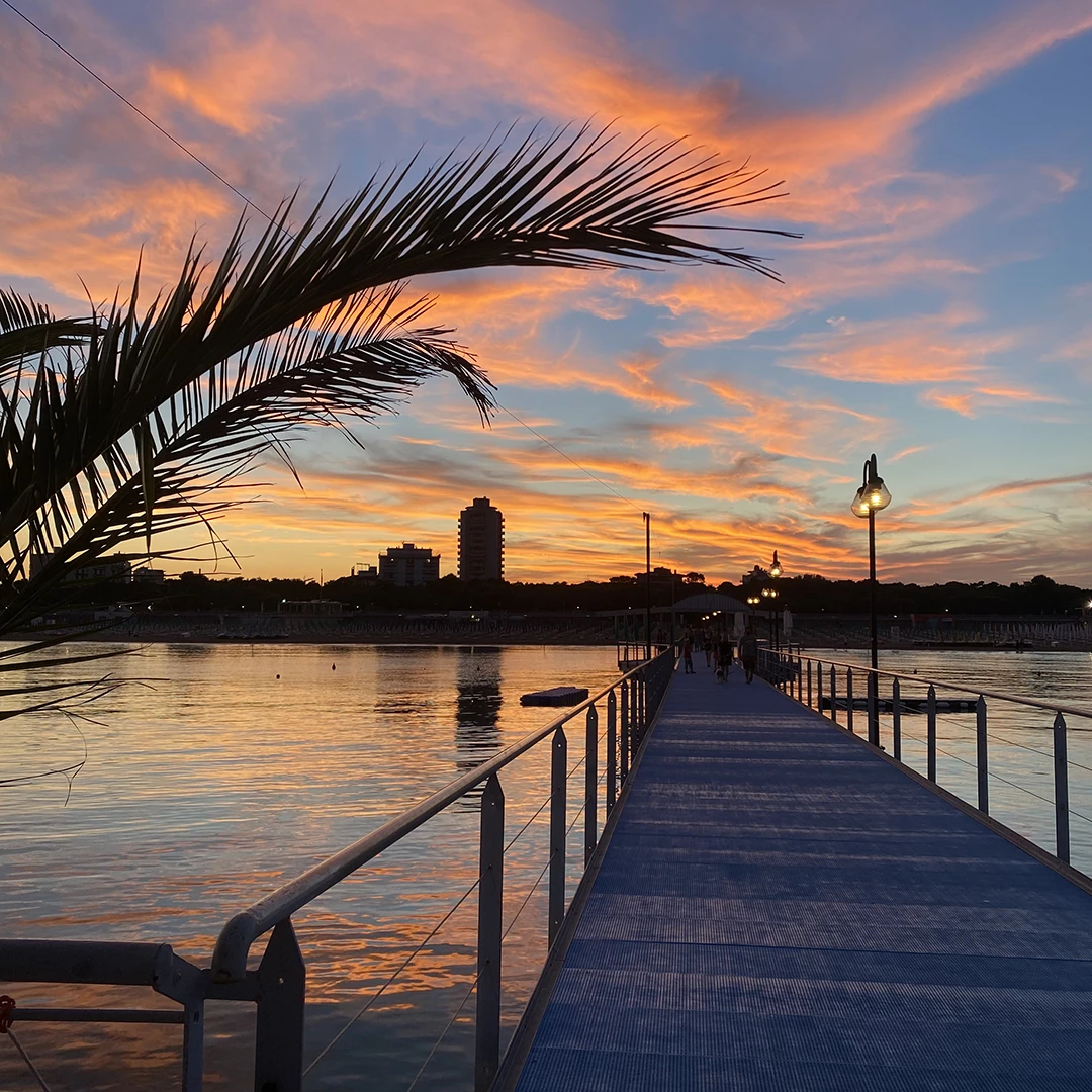 Si vede un tramonto ripreso dal punto di vista di chi sta facendo un aperitivo dalla Pagoda. Si vedono gli avvolgenti colori del cielo e lo skyline di Lignano Pineta